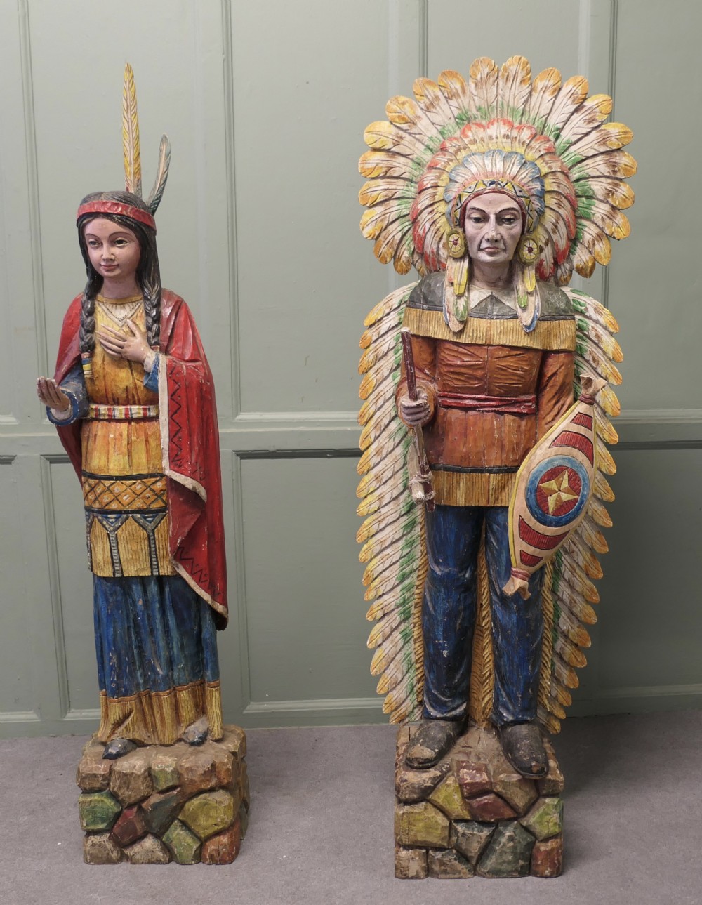 A pair of hand-carved, polychrome painted wooden statues depicting figures in traditional Native American attire set on stone-effect bases.