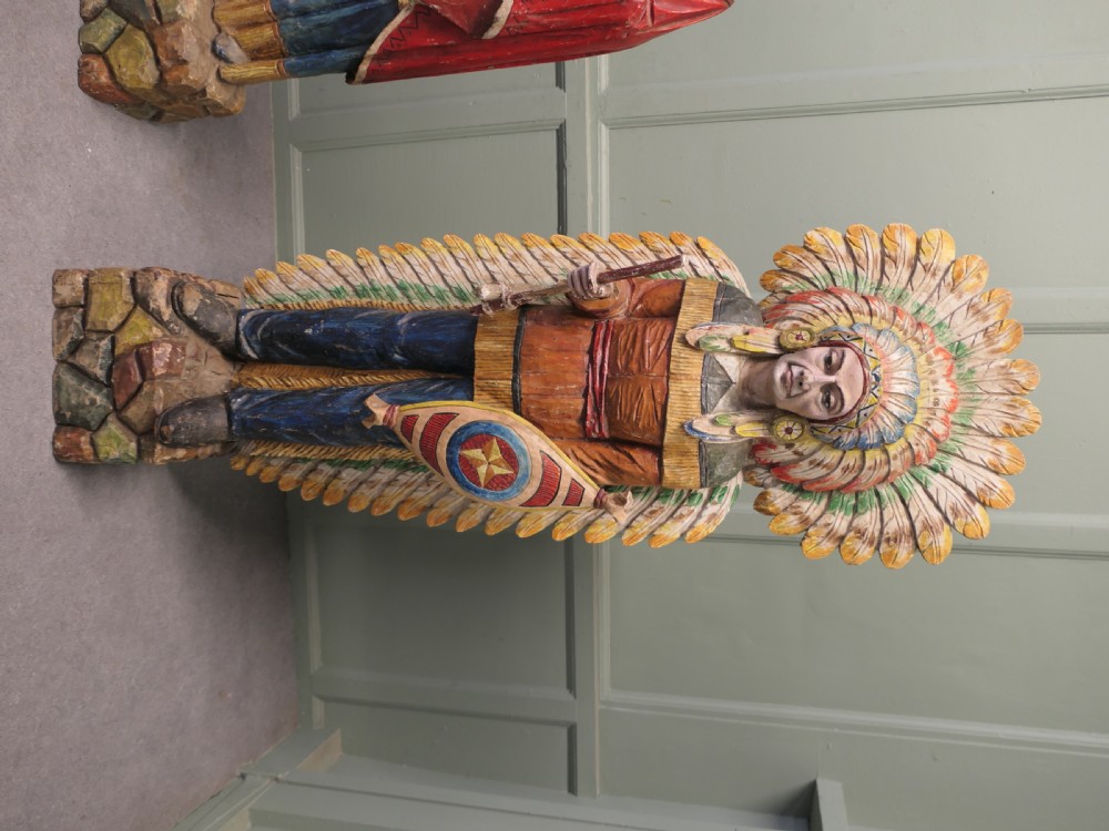 A pair of hand-carved, polychrome painted wooden statues depicting figures in traditional Native American attire set on stone-effect bases. View II