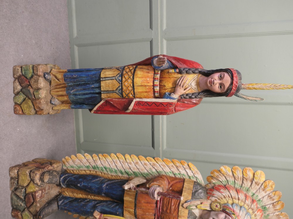 A pair of hand-carved, polychrome painted wooden statues depicting figures in traditional Native American attire set on stone-effect bases. View III