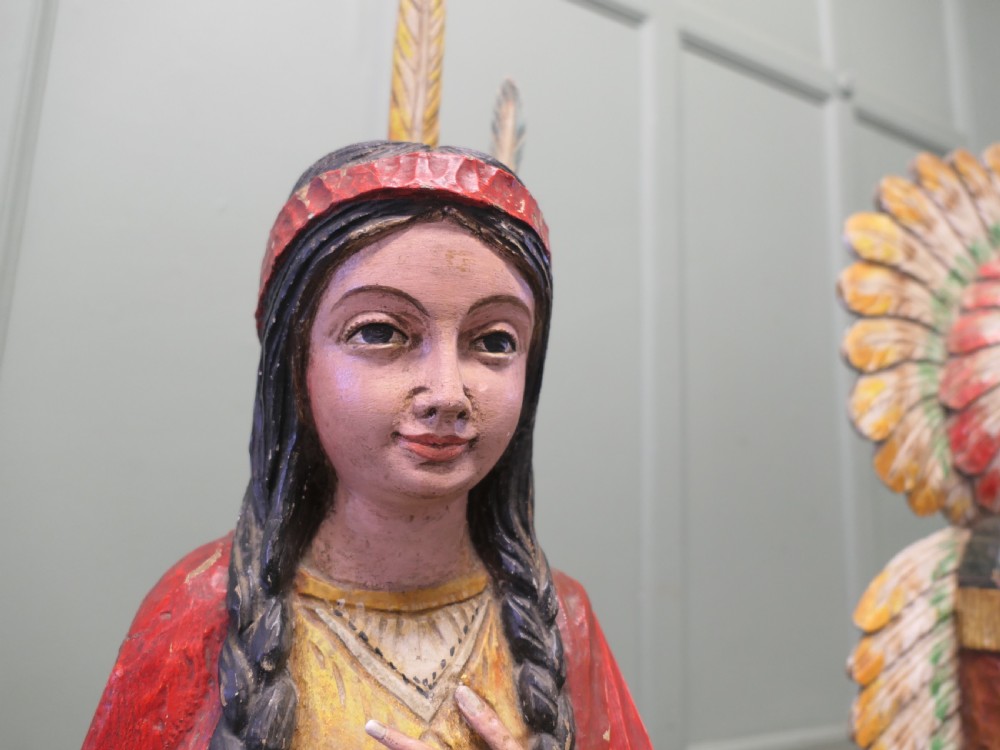 A pair of hand-carved, polychrome painted wooden statues depicting figures in traditional Native American attire set on stone-effect bases. View IV
