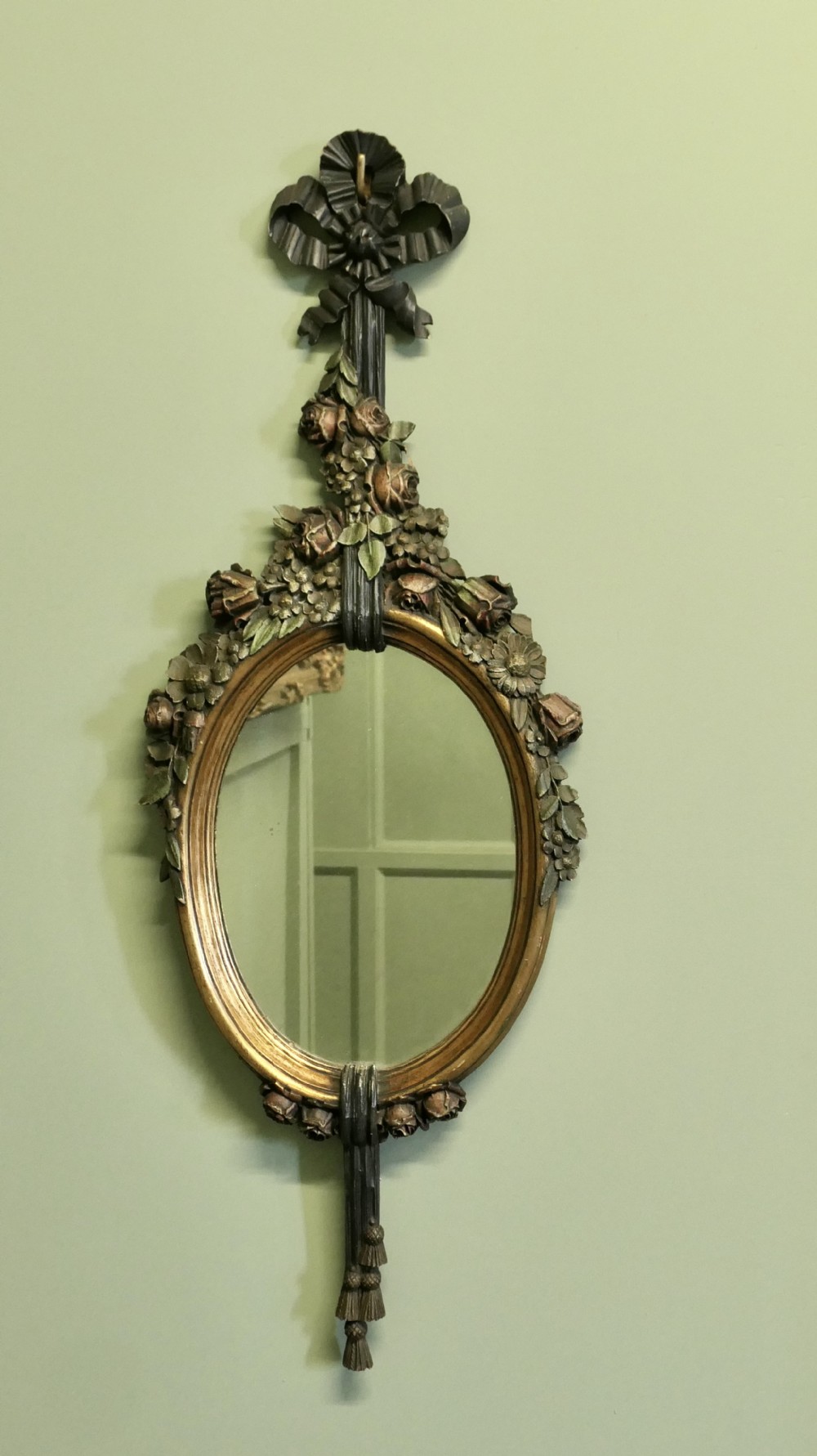 A vertically oriented oval French mirror featuring a carved ribbon bow crest, polychrome painted floral garlands, and tassel drop detailing at the base.