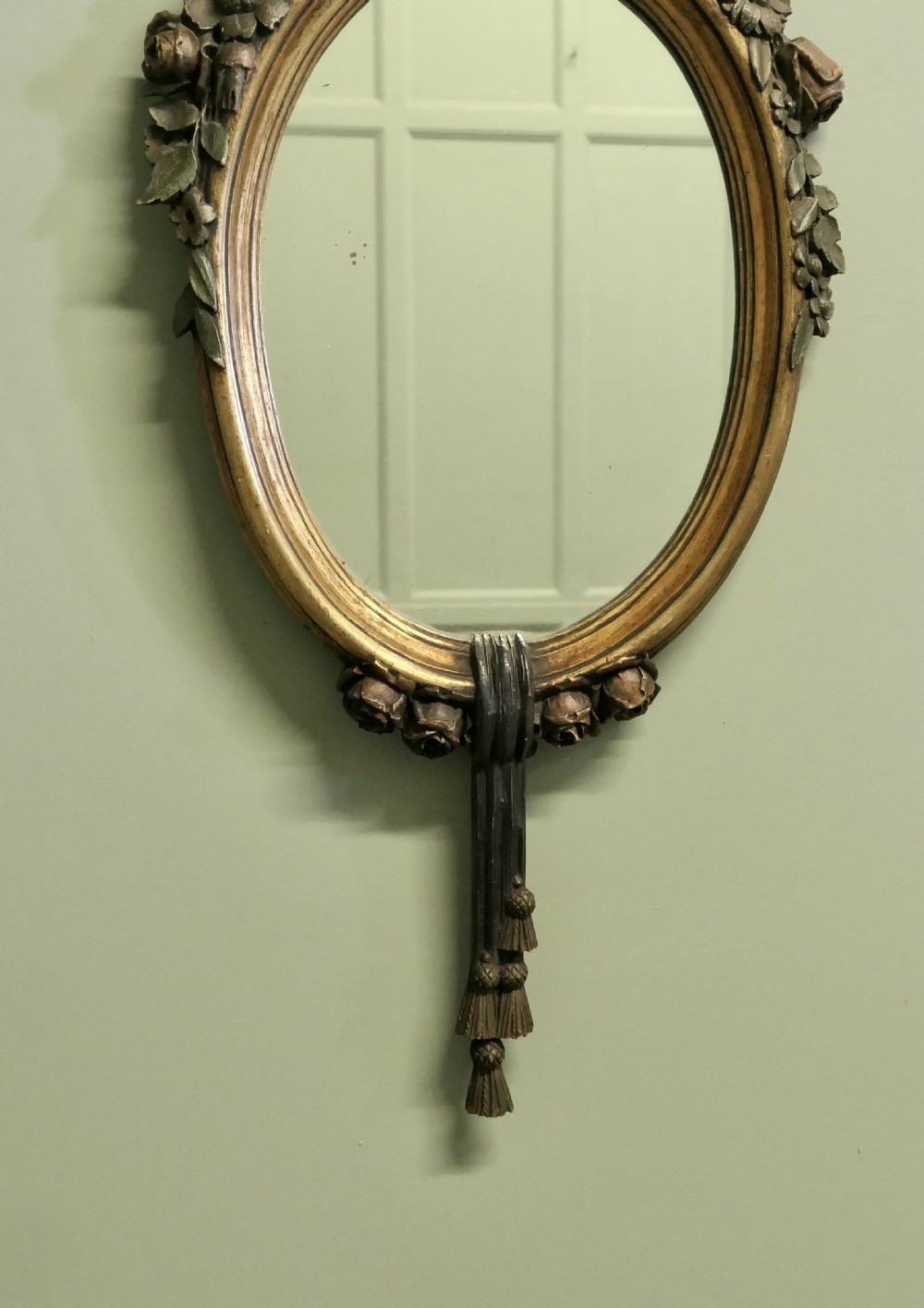 A vertically oriented oval French mirror featuring a carved ribbon bow crest, polychrome painted floral garlands, and tassel drop detailing at the base. View III