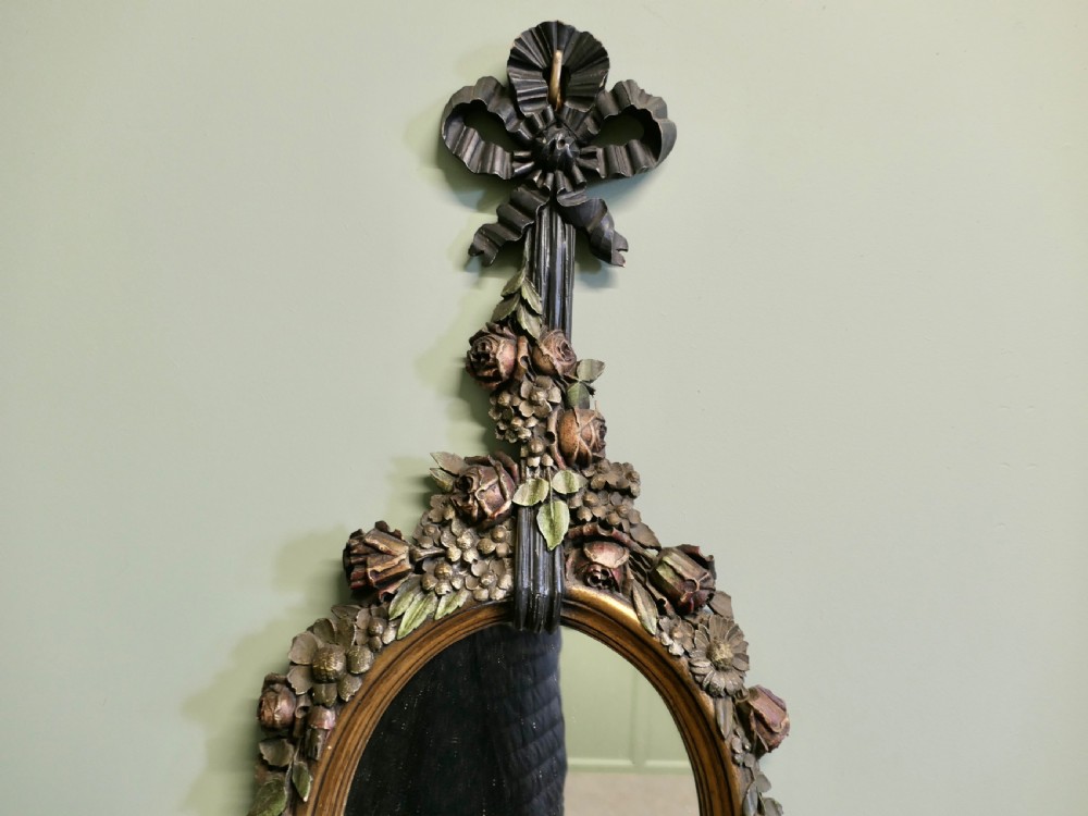 A vertically oriented oval French mirror featuring a carved ribbon bow crest, polychrome painted floral garlands, and tassel drop detailing at the base. View IV