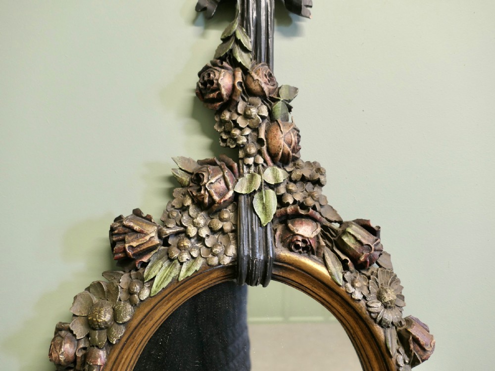 A vertically oriented oval French mirror featuring a carved ribbon bow crest, polychrome painted floral garlands, and tassel drop detailing at the base. View VI