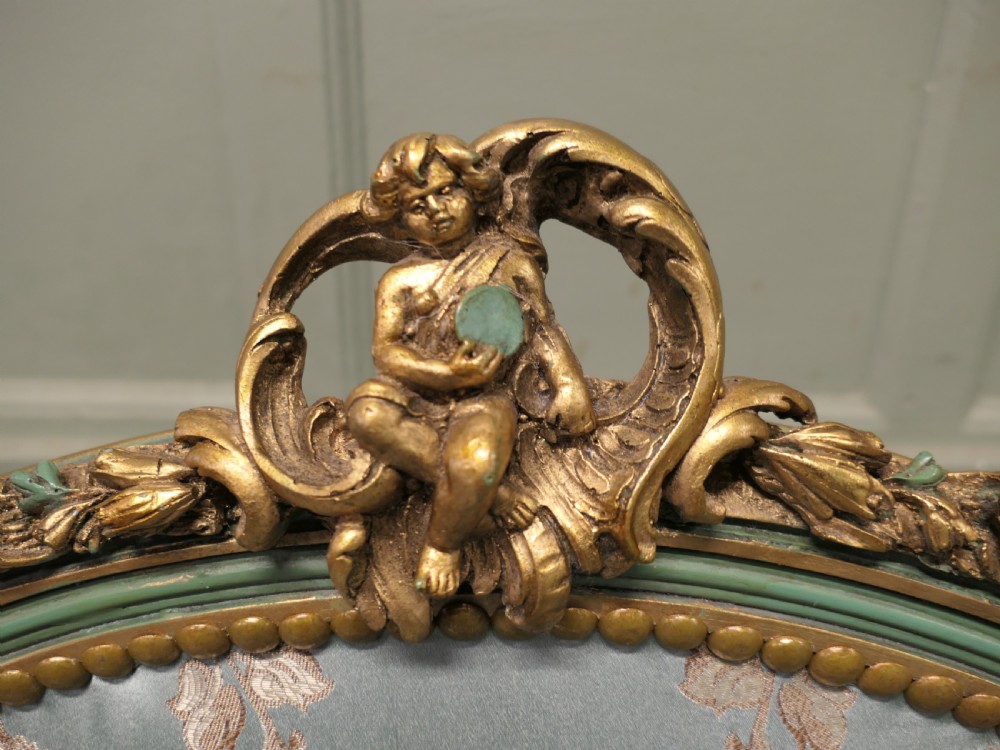 A French Rococo style settee with a green and gold parcel-gilt wooden frame, carved putti cresting, and light green floral silk upholstery. View V