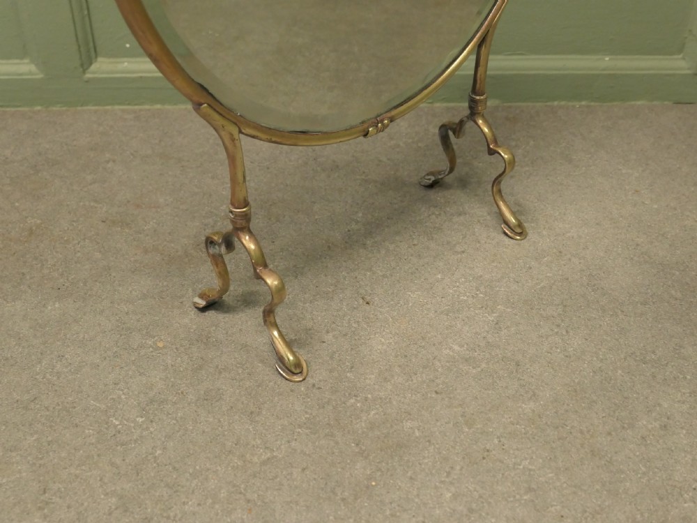An Art Nouveau brass fire screen featuring a circular mirror frame topped with an intricate whiplash crest and resting on two curved metal legs. View II