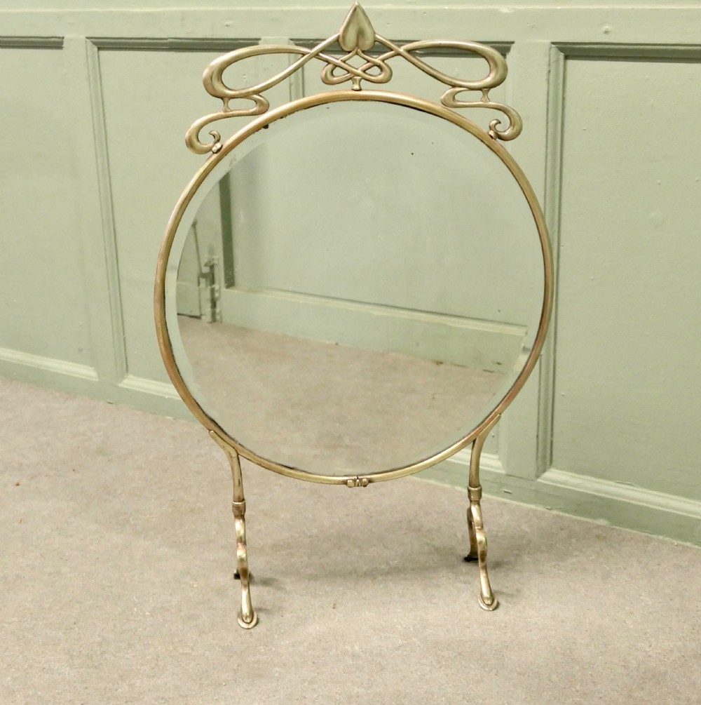 An Art Nouveau brass fire screen featuring a circular mirror frame topped with an intricate whiplash crest and resting on two curved metal legs.