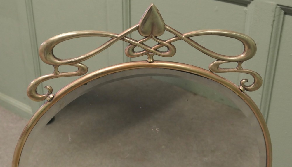 An Art Nouveau brass fire screen featuring a circular mirror frame topped with an intricate whiplash crest and resting on two curved metal legs. View IV