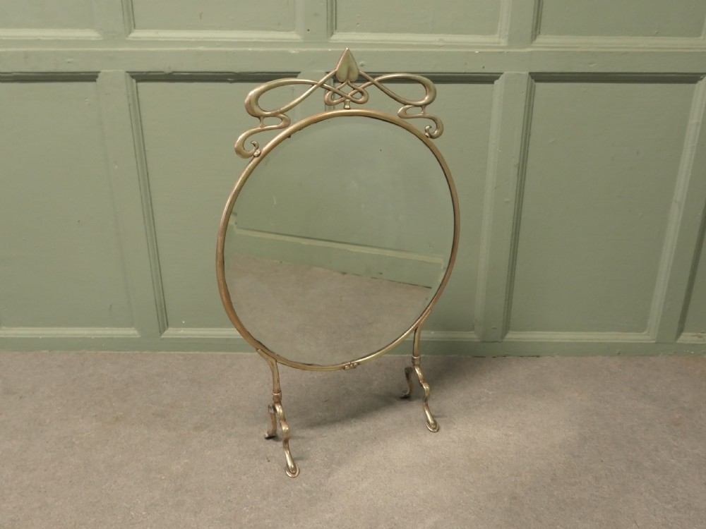 An Art Nouveau brass fire screen featuring a circular mirror frame topped with an intricate whiplash crest and resting on two curved metal legs. View V