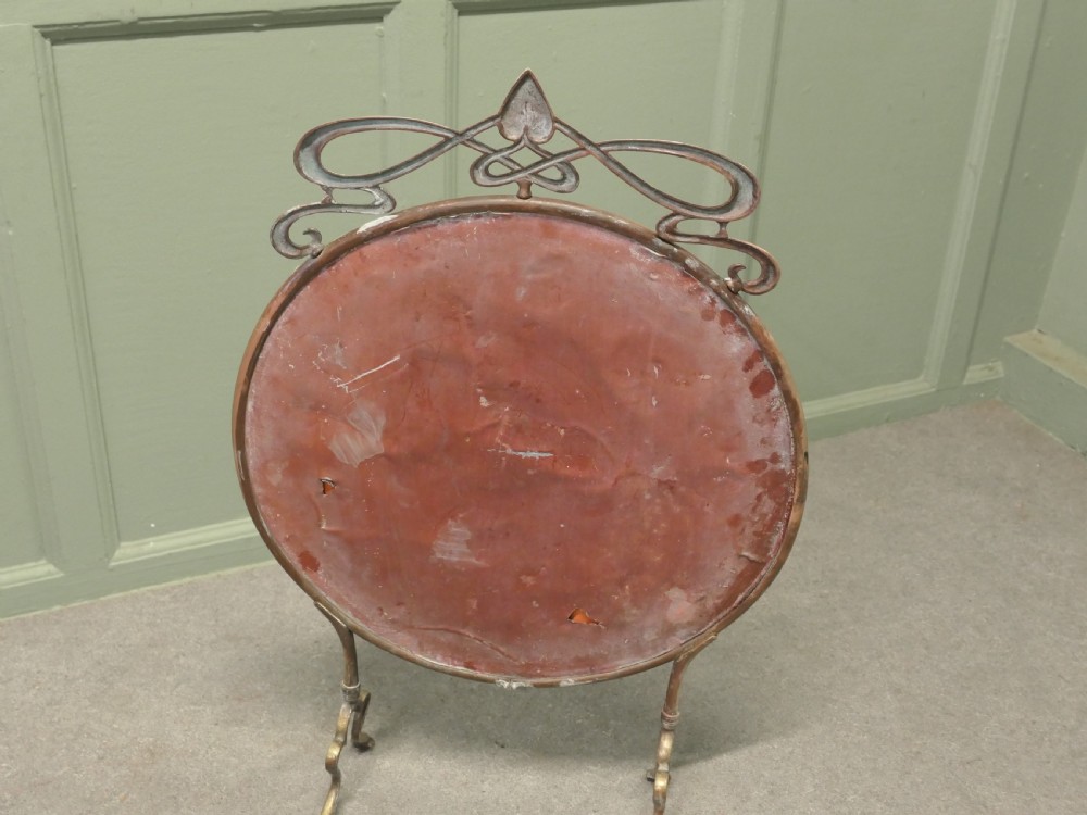 An Art Nouveau brass fire screen featuring a circular mirror frame topped with an intricate whiplash crest and resting on two curved metal legs. View VI