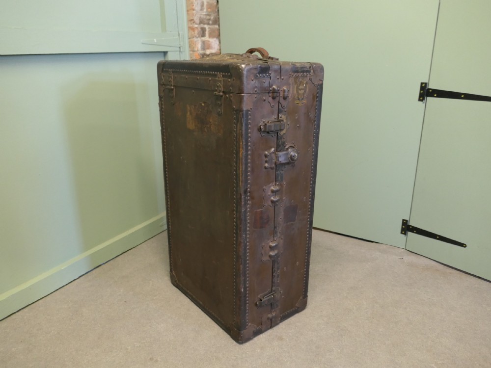 An upright dark brown metal-bound fitted steamer trunk wardrobe featuring riveted corners and heavy brass latches.