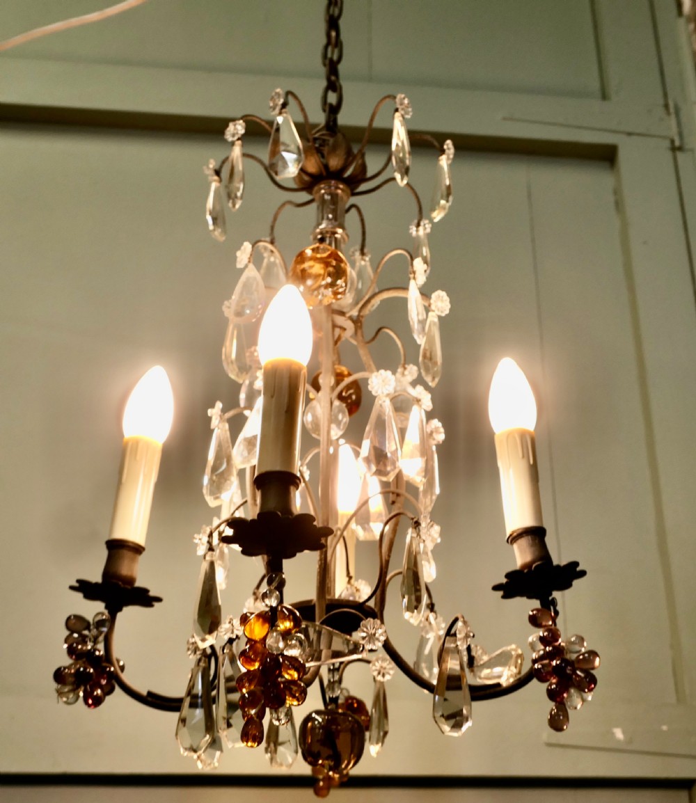 A French metal chandelier featuring scroll arms, electric candle sleeves, clear crystal droplets, and coloured glass fruit ornaments in amber and mauve tones. View IV