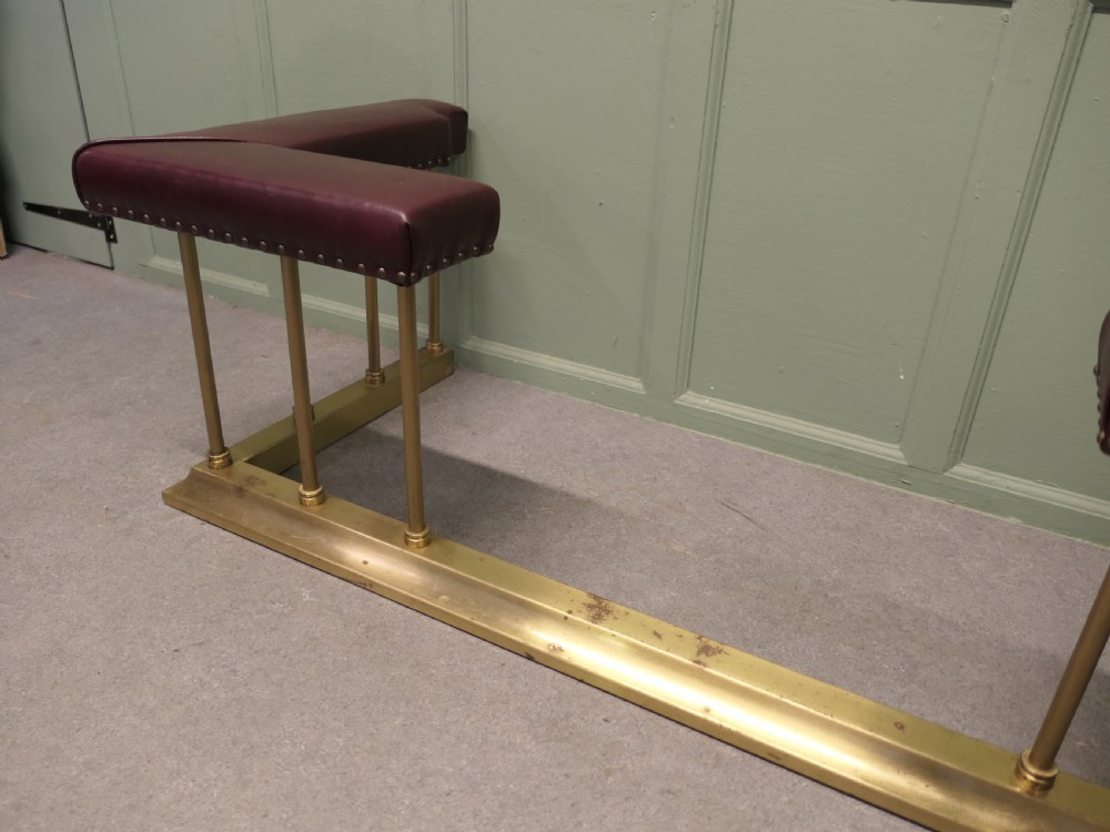An Art Deco brass club fender featuring corner seats upholstered in burgundy leather with brass stud trimming, supported by vertical brass columns.