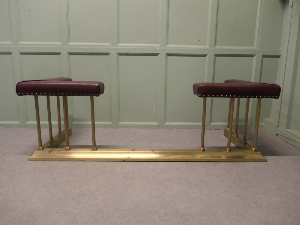 An Art Deco brass club fender featuring corner seats upholstered in burgundy leather with brass stud trimming, supported by vertical brass columns. View II