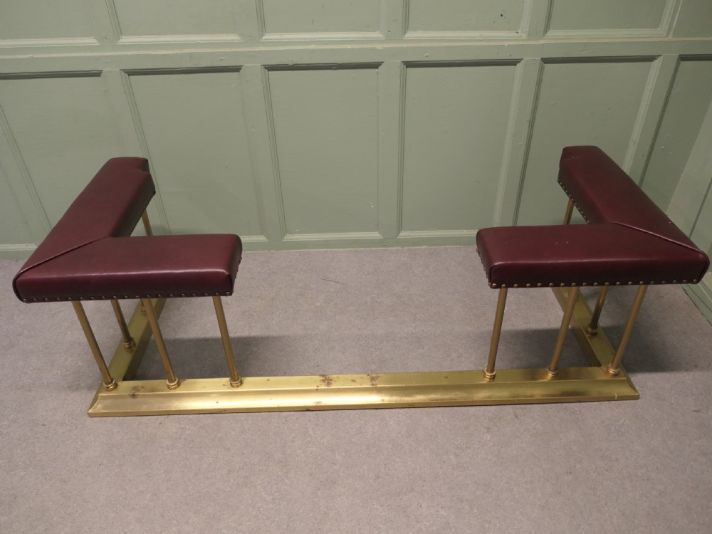 An Art Deco brass club fender featuring corner seats upholstered in burgundy leather with brass stud trimming, supported by vertical brass columns. View V