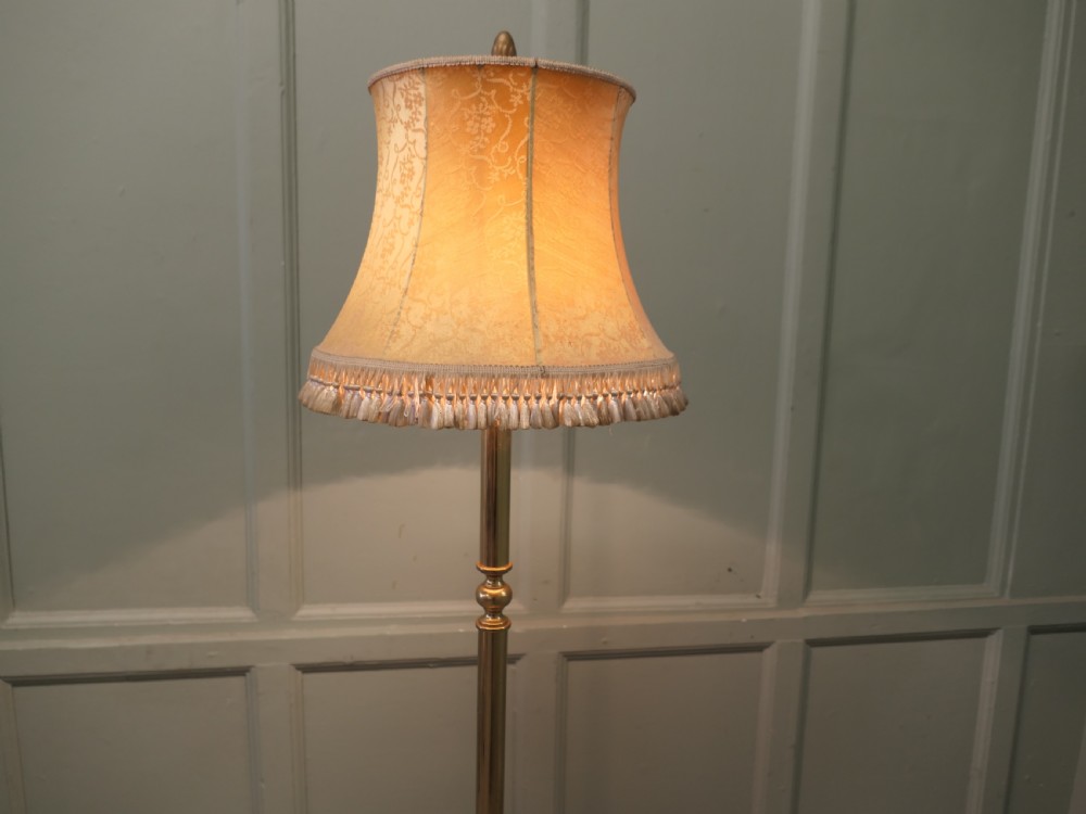 A slender turned brass floor lamp with a moulded circular base, topped with a cream patterned fabric shade featuring fringe trim. View IV