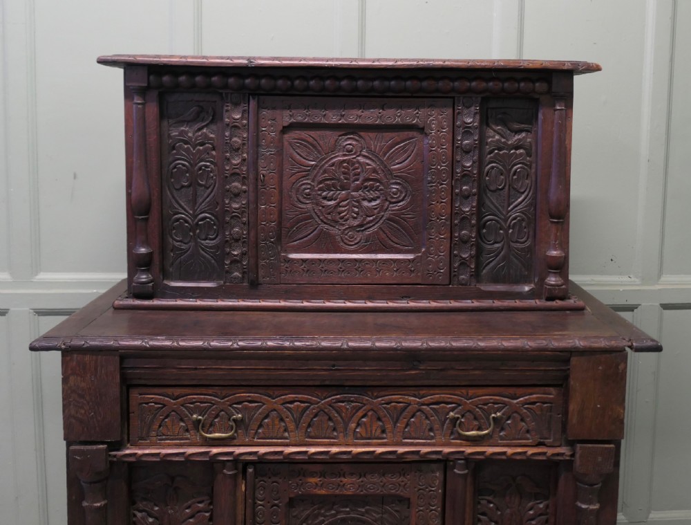 A two-tier dark carved oak court cupboard featuring relief panels with carved foliage, turned side columns, a upper cupboard door, and a lower drawer over a central cabinet door. View II