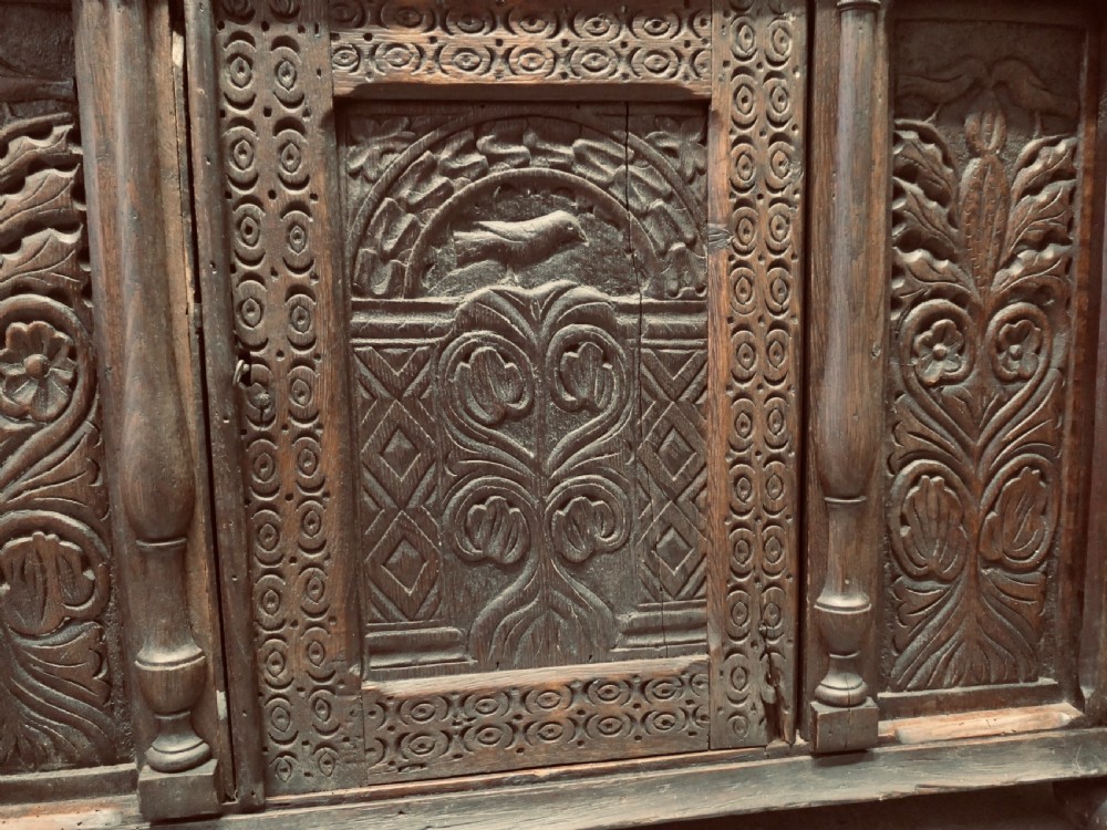 A two-tier dark carved oak court cupboard featuring relief panels with carved foliage, turned side columns, a upper cupboard door, and a lower drawer over a central cabinet door. View III