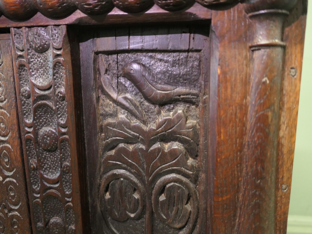 A two-tier dark carved oak court cupboard featuring relief panels with carved foliage, turned side columns, a upper cupboard door, and a lower drawer over a central cabinet door. View IV