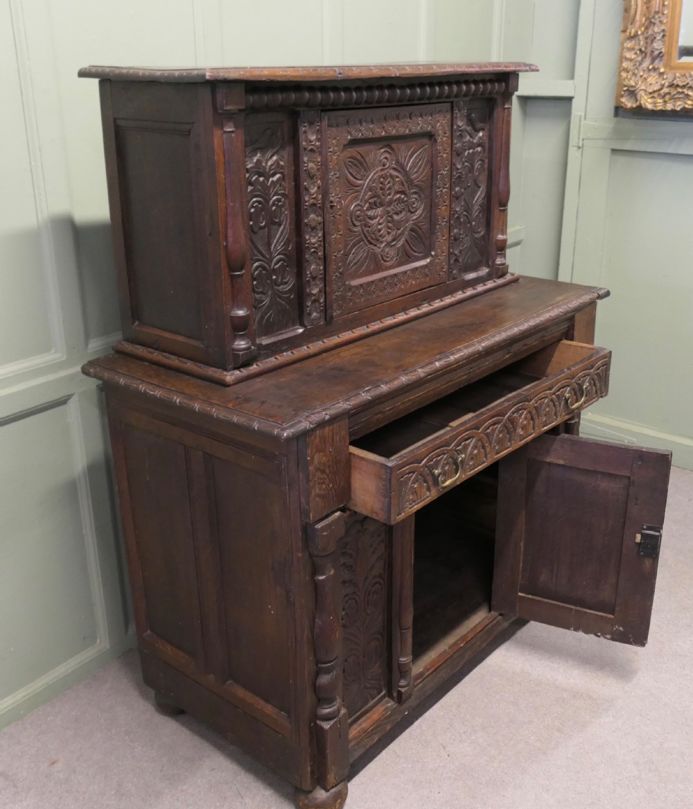 A two-tier dark carved oak court cupboard featuring relief panels with carved foliage, turned side columns, a upper cupboard door, and a lower drawer over a central cabinet door. View V