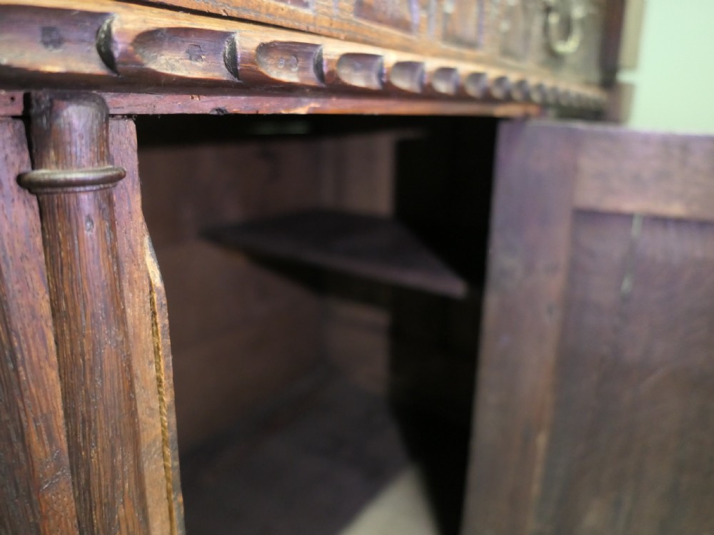 A two-tier dark carved oak court cupboard featuring relief panels with carved foliage, turned side columns, a upper cupboard door, and a lower drawer over a central cabinet door. View VI
