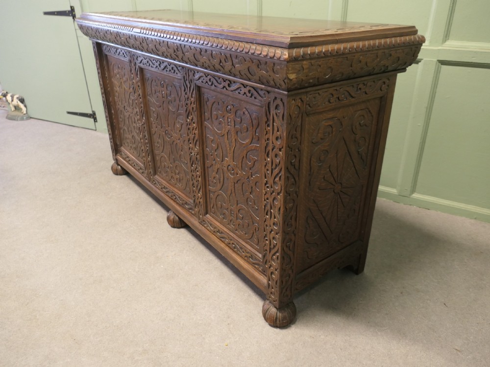 A solid golden oak three-door cupboard featuring intricate relief carving across the doors and frame, supported by carved melon-bulb feet.