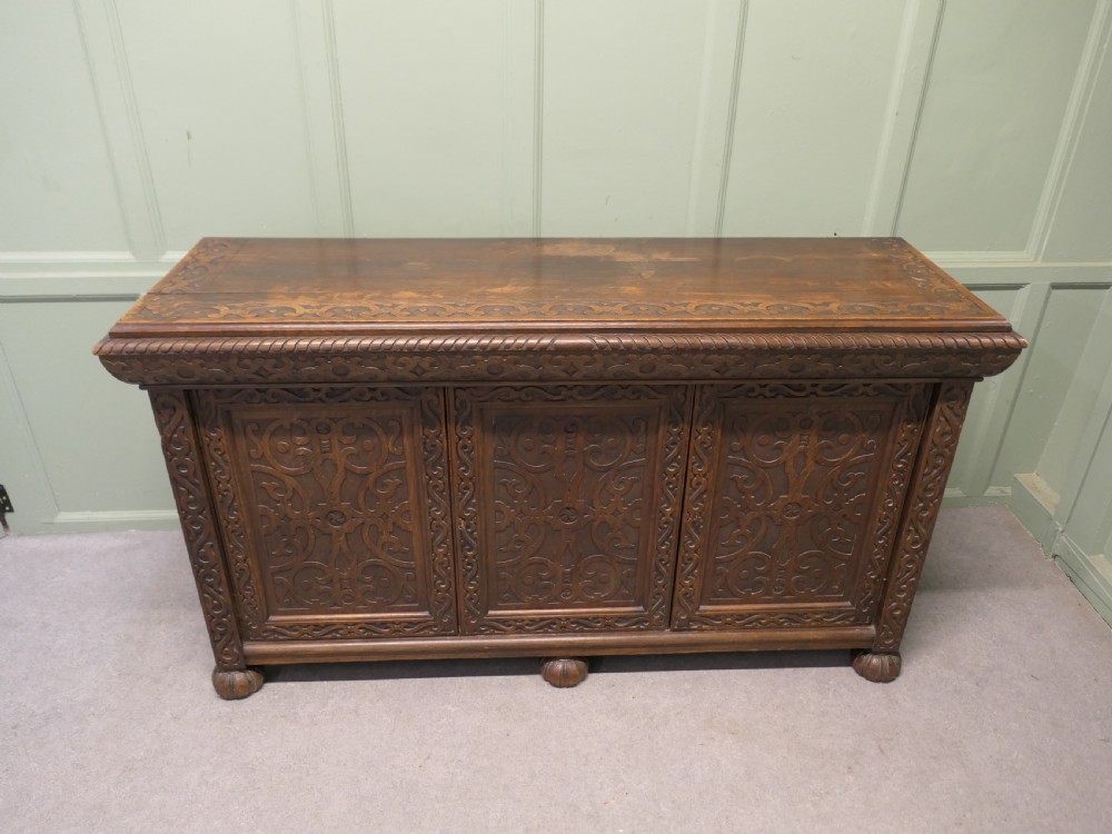 A solid golden oak three-door cupboard featuring intricate relief carving across the doors and frame, supported by carved melon-bulb feet. View II