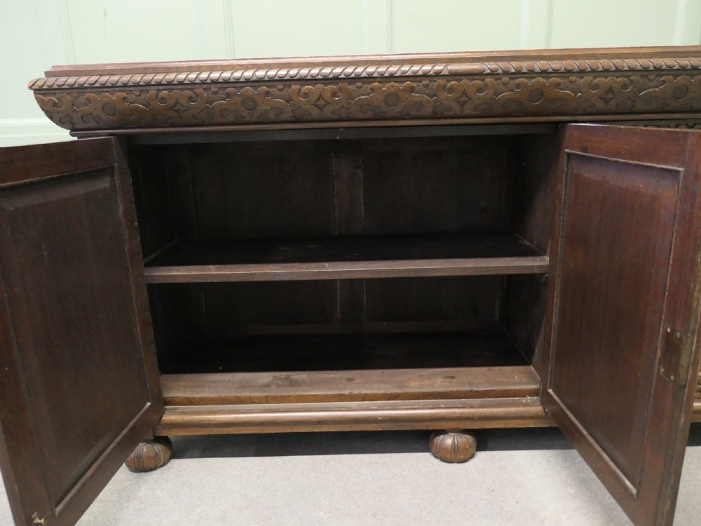 A solid golden oak three-door cupboard featuring intricate relief carving across the doors and frame, supported by carved melon-bulb feet. View III