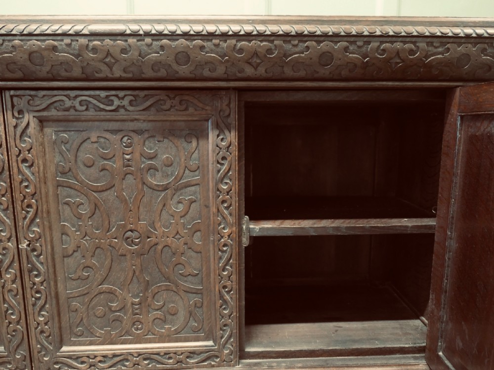 A solid golden oak three-door cupboard featuring intricate relief carving across the doors and frame, supported by carved melon-bulb feet. View IV