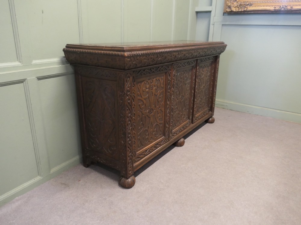 A solid golden oak three-door cupboard featuring intricate relief carving across the doors and frame, supported by carved melon-bulb feet. View V