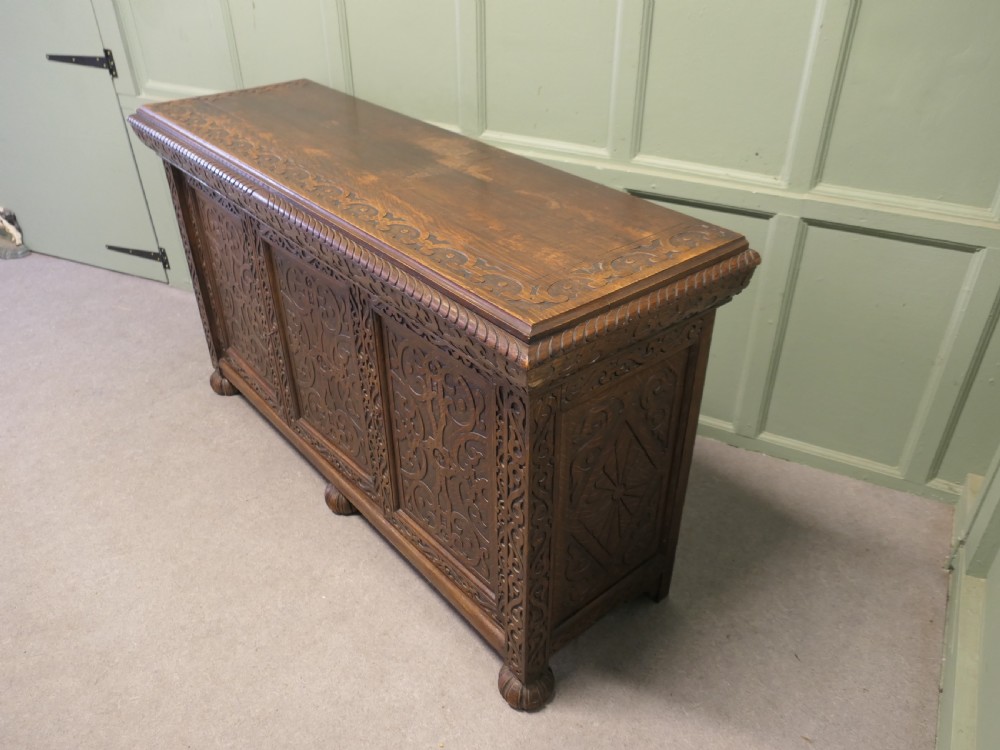 A solid golden oak three-door cupboard featuring intricate relief carving across the doors and frame, supported by carved melon-bulb feet. View VI