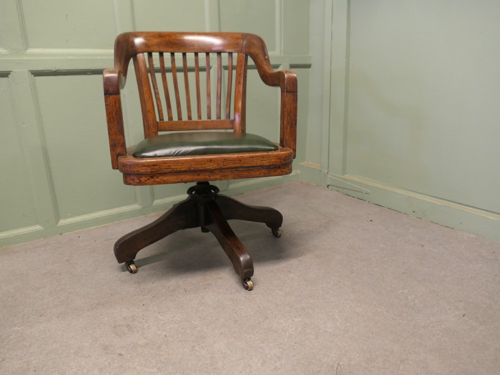 An Art Deco oak swivel desk chair featuring a curved backrest with vertical slats, a green leather padded seat, and a quadruped base on castors.