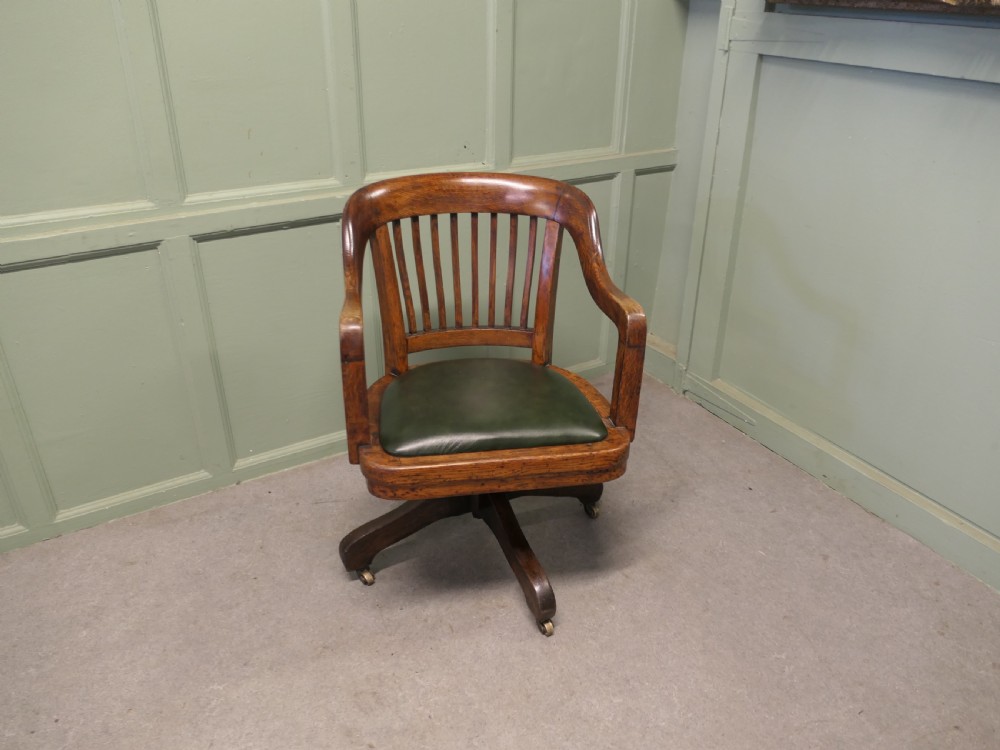 An Art Deco oak swivel desk chair featuring a curved backrest with vertical slats, a green leather padded seat, and a quadruped base on castors. View II