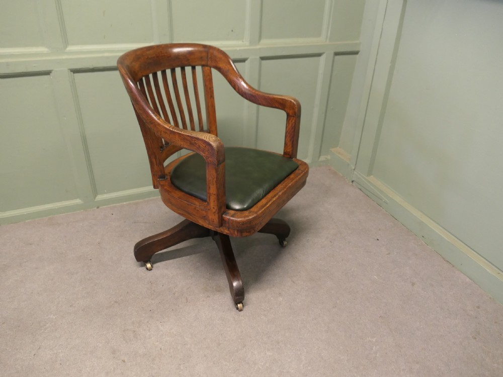 An Art Deco oak swivel desk chair featuring a curved backrest with vertical slats, a green leather padded seat, and a quadruped base on castors. View III