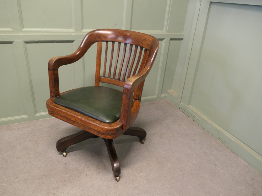 An Art Deco oak swivel desk chair featuring a curved backrest with vertical slats, a green leather padded seat, and a quadruped base on castors. View V