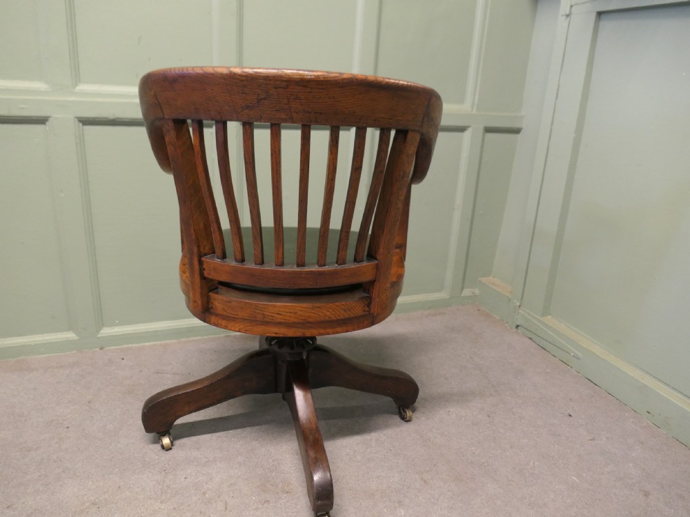 An Art Deco oak swivel desk chair featuring a curved backrest with vertical slats, a green leather padded seat, and a quadruped base on castors. View VI