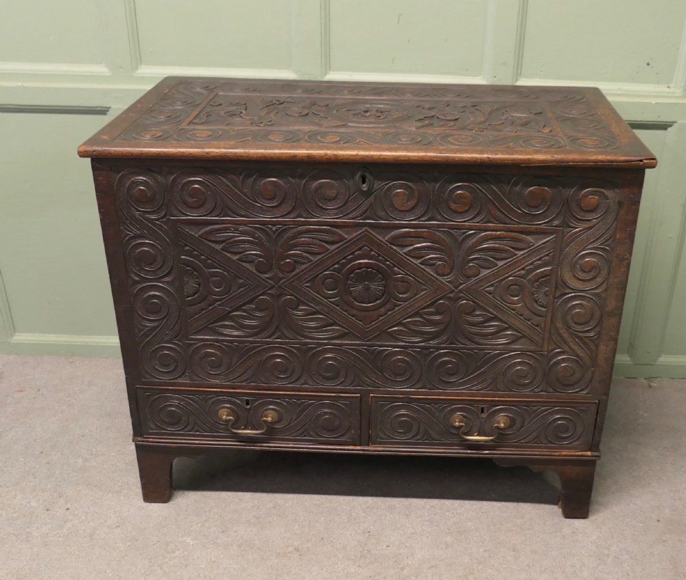 A dark carved oak mule chest featuring a hinged lid, relief-carved scrollwork and geometric patterns, and two drawers at the bottom with brass drop handles.