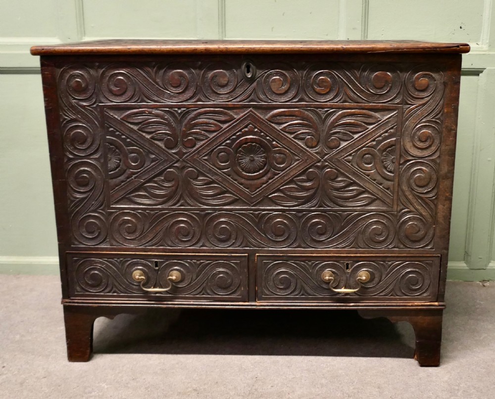 A dark carved oak mule chest featuring a hinged lid, relief-carved scrollwork and geometric patterns, and two drawers at the bottom with brass drop handles. View II