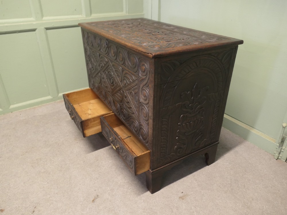 A dark carved oak mule chest featuring a hinged lid, relief-carved scrollwork and geometric patterns, and two drawers at the bottom with brass drop handles. View VI