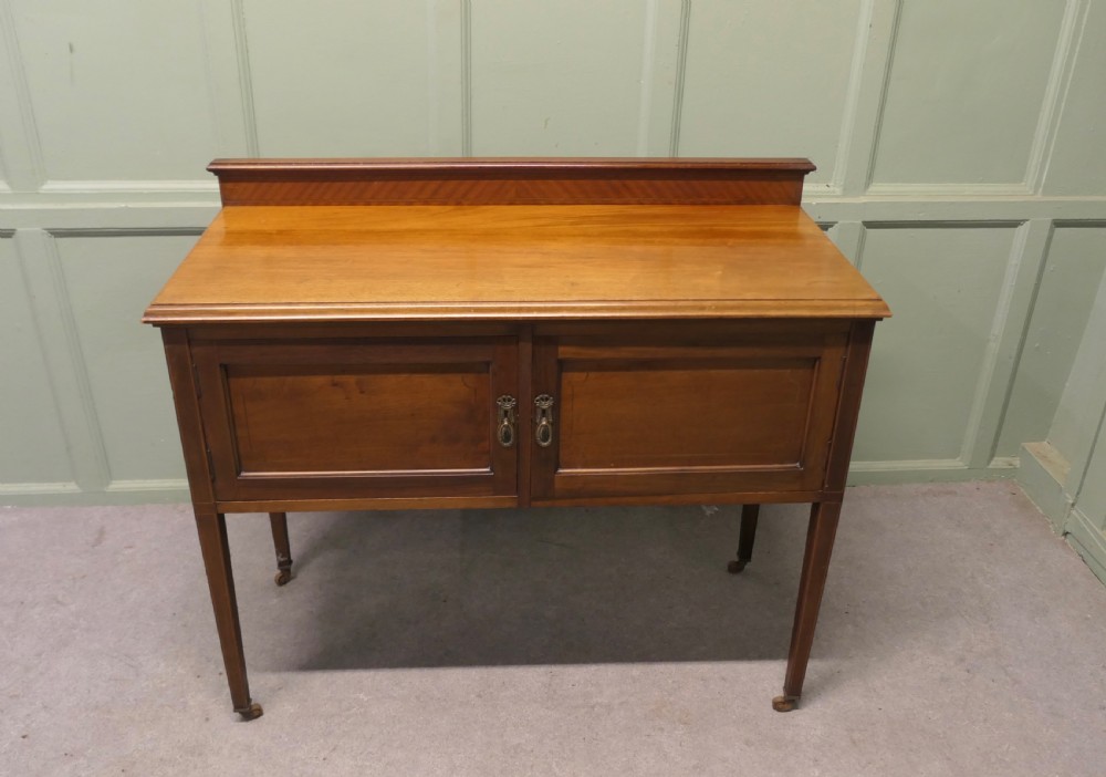 A late Victorian warm brown walnut side cupboard featuring twin panelled doors, a low gallery back with parquetry inlay, and square tapered legs on brass castors.