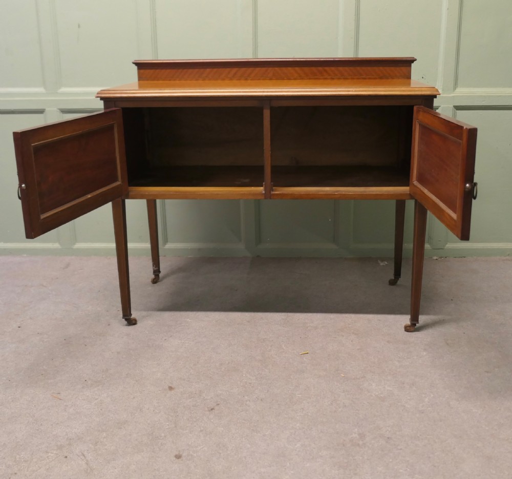 A late Victorian warm brown walnut side cupboard featuring twin panelled doors, a low gallery back with parquetry inlay, and square tapered legs on brass castors. View II