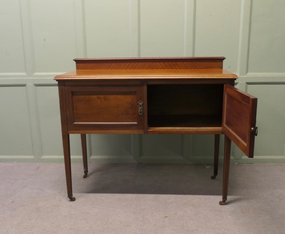 A late Victorian warm brown walnut side cupboard featuring twin panelled doors, a low gallery back with parquetry inlay, and square tapered legs on brass castors. View V