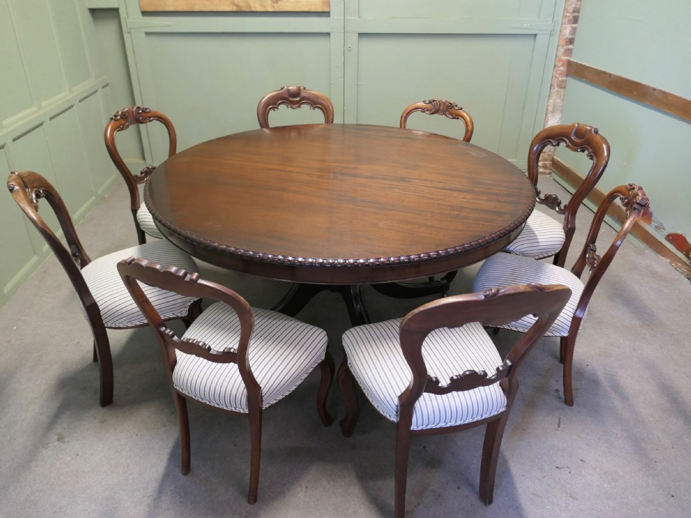 A circular dark mahogany dining table surrounded by eight matching carved balloon-back dining chairs with blue and white striped fabric seats.