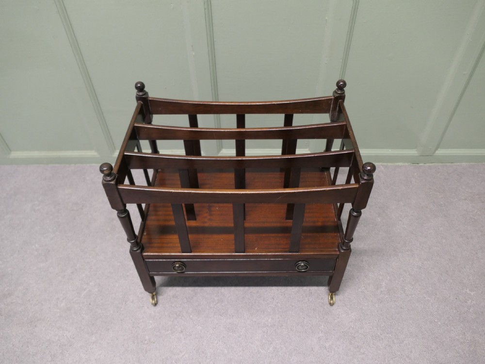 A dark walnut Canterbury magazine stand featuring slatted divisions, turned corner posts with finials, a lower drawer, and brass castors.