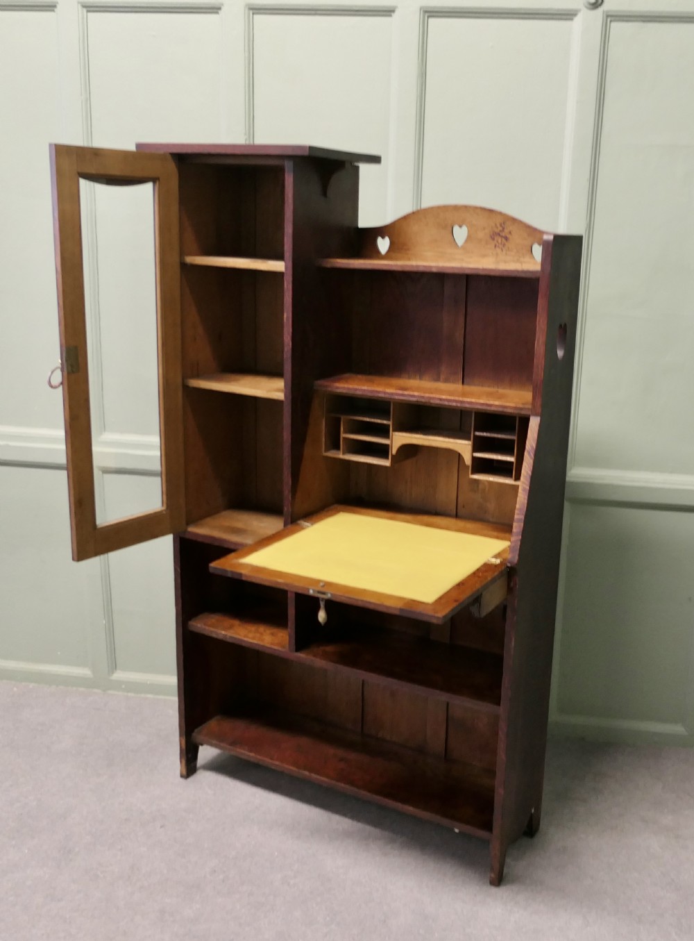 An Arts and Crafts golden oak student bureau bookcase featuring a glazed door cabinet, heart cut-outs, a drop-front writing slope, and open storage shelves. View III
