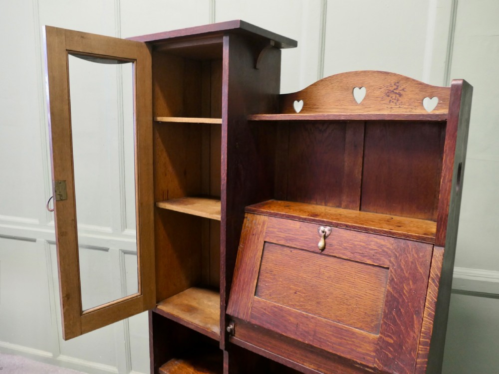 An Arts and Crafts golden oak student bureau bookcase featuring a glazed door cabinet, heart cut-outs, a drop-front writing slope, and open storage shelves. View IV