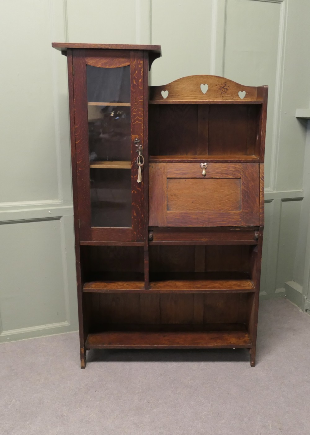 An Arts and Crafts golden oak student bureau bookcase featuring a glazed door cabinet, heart cut-outs, a drop-front writing slope, and open storage shelves. View V