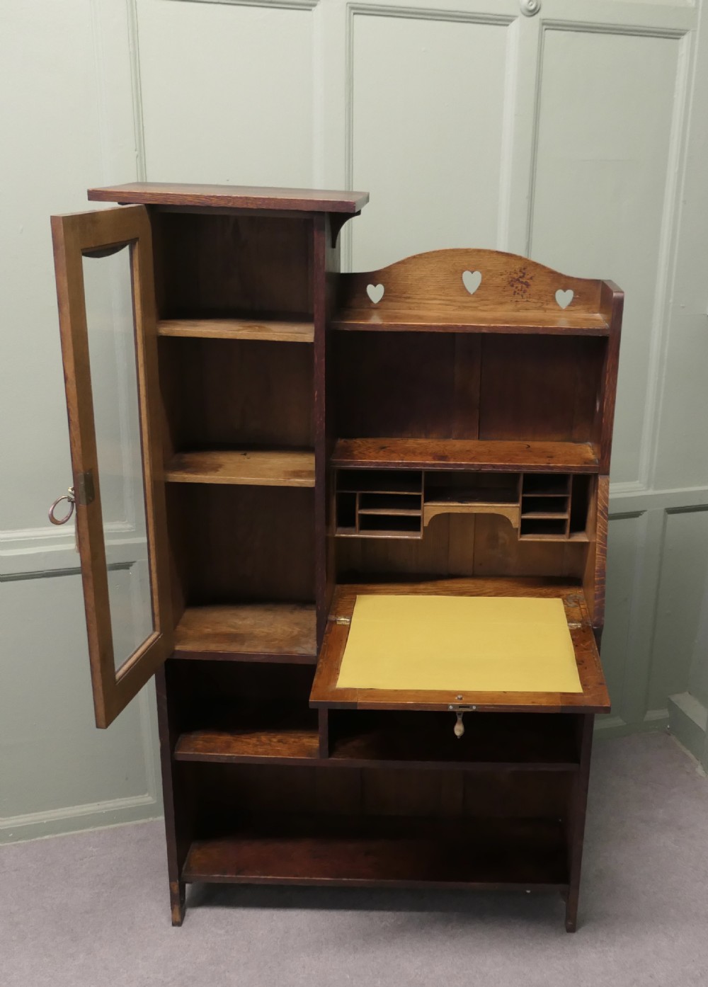 An Arts and Crafts golden oak student bureau bookcase featuring a glazed door cabinet, heart cut-outs, a drop-front writing slope, and open storage shelves. View VI