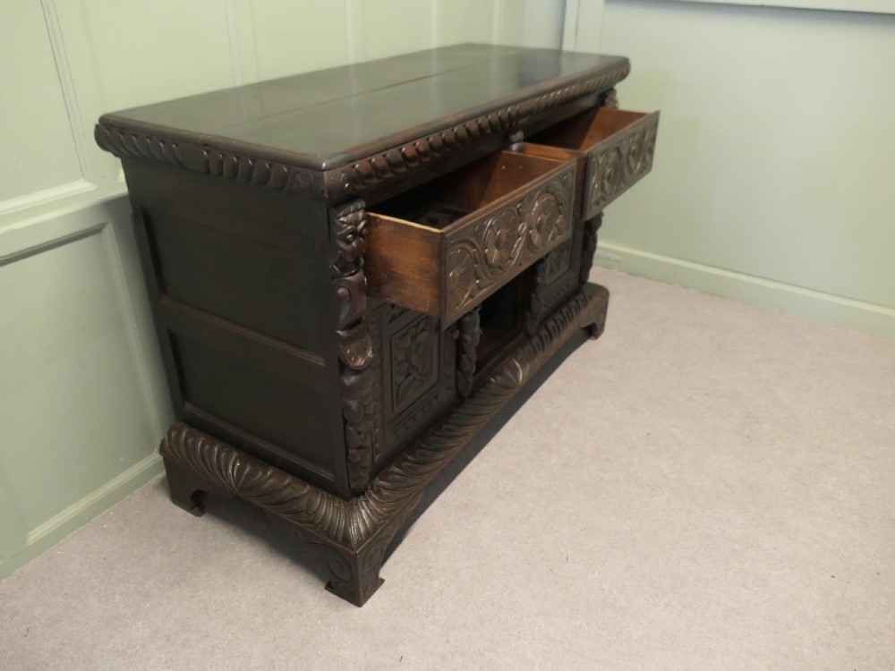 A dark carved oak dresser base featuring Green Man masks, floral panels, and gadrooned borders. View V
