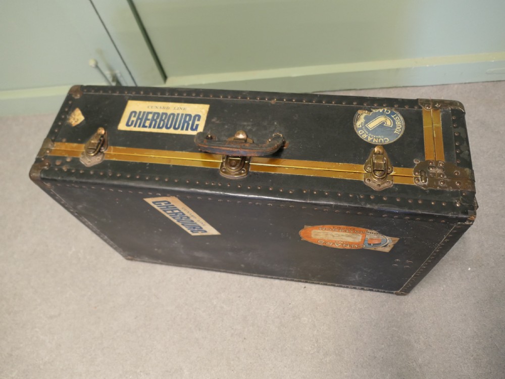 A black canvas cabin trunk featuring brass latches, riveted edges, a central handle, and original Cunard Line Cherbourg travel labels.